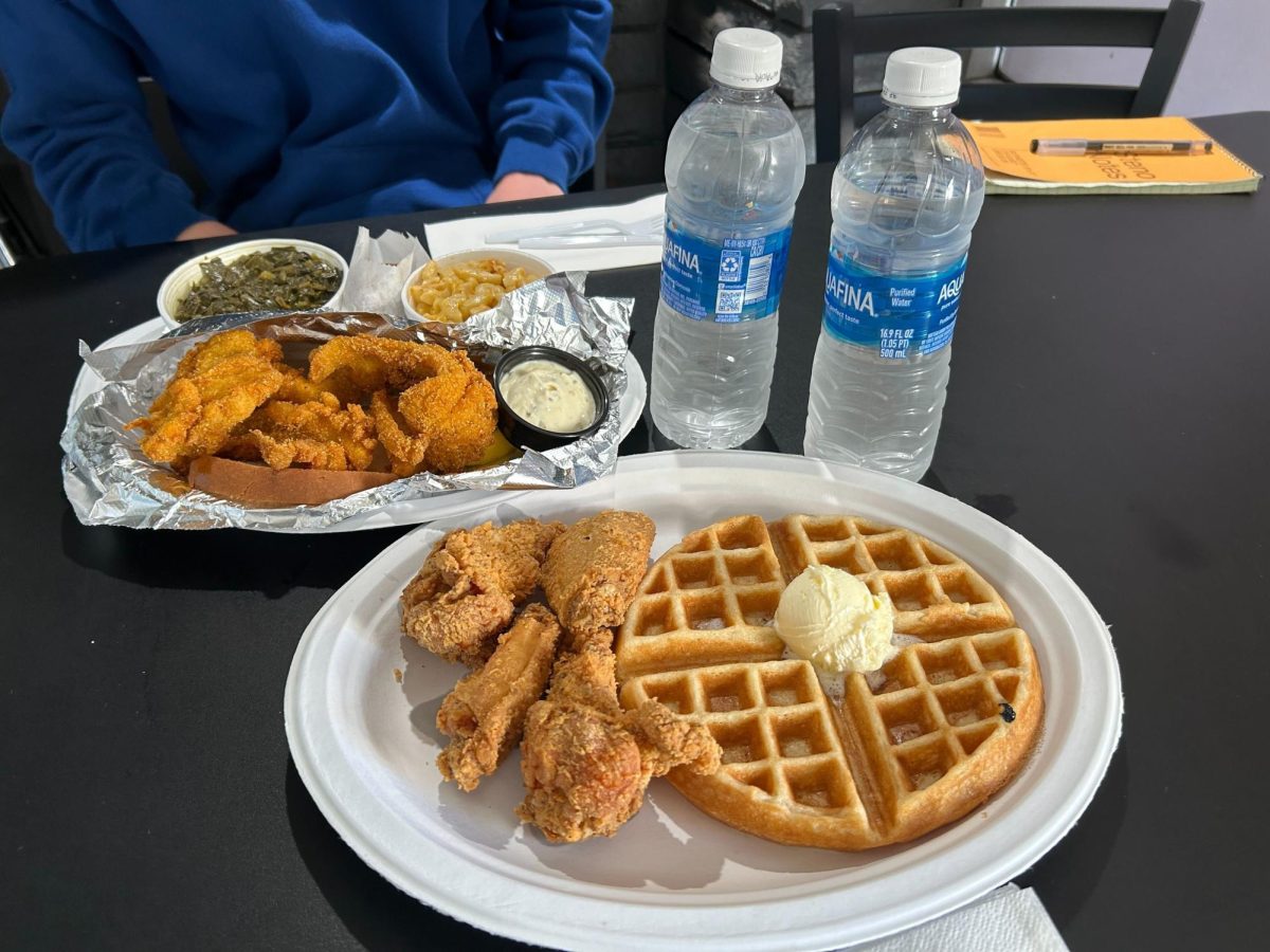 A plate stacked with four crispy, golden fried chicken pieces and four fluffy waffles is topped with butter, a sprinkle of sugar and a drizzle of rich maple syrup. 